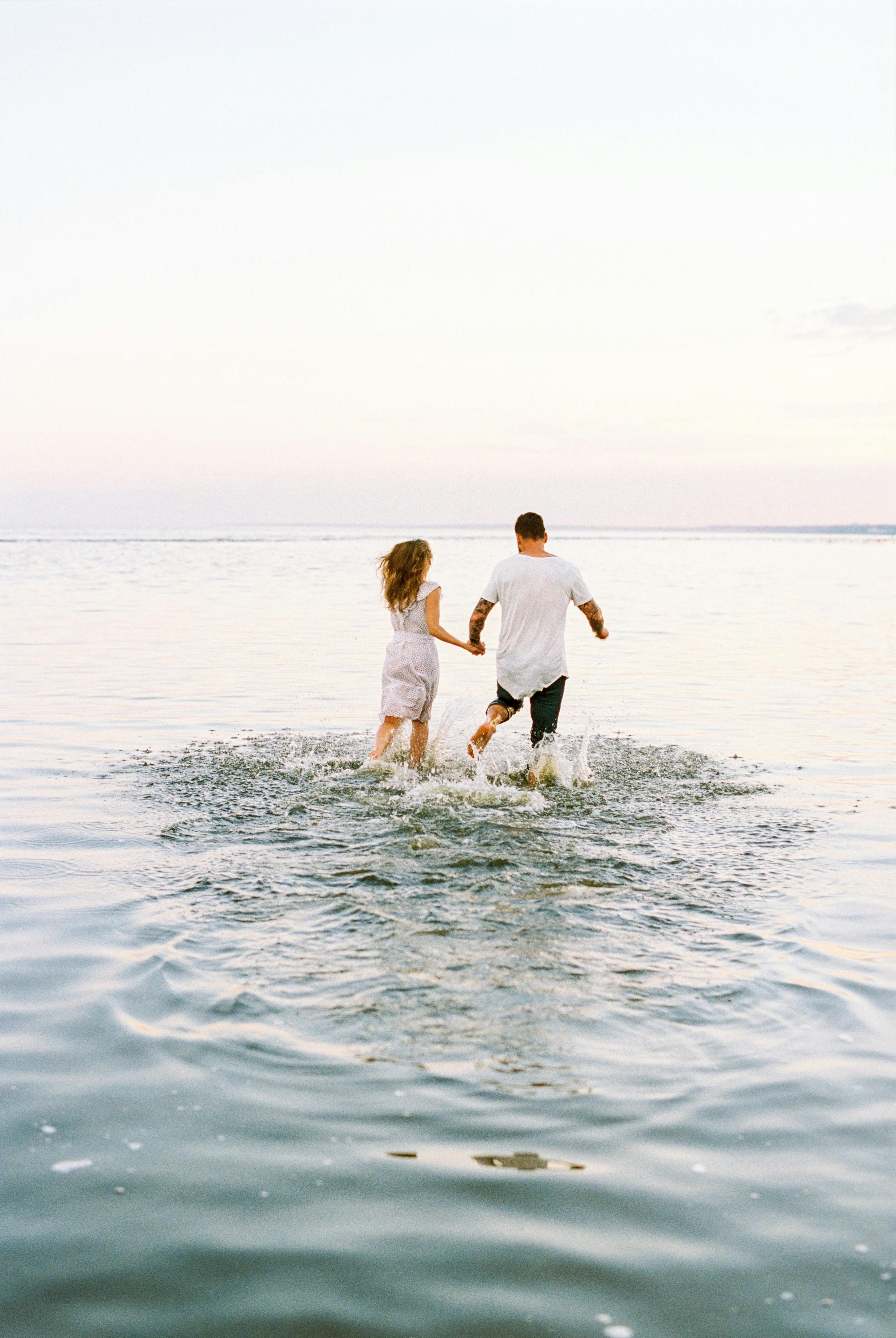 A young couple holding hands and running into a tranquil lake, splashing water around, during a peaceful evening.