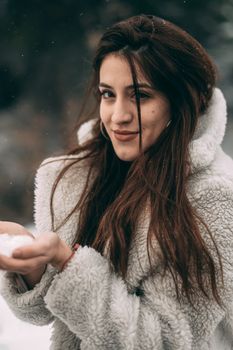 A brunette woman in a white coat holding snow, enjoying a winter day.