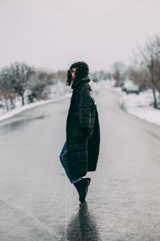 Fashionably dressed woman in black coat walks down a snowy road during winter.
