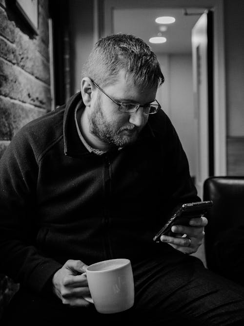 A Man Sitting on a Chair while Holding a Ceramic Mug and His Phone