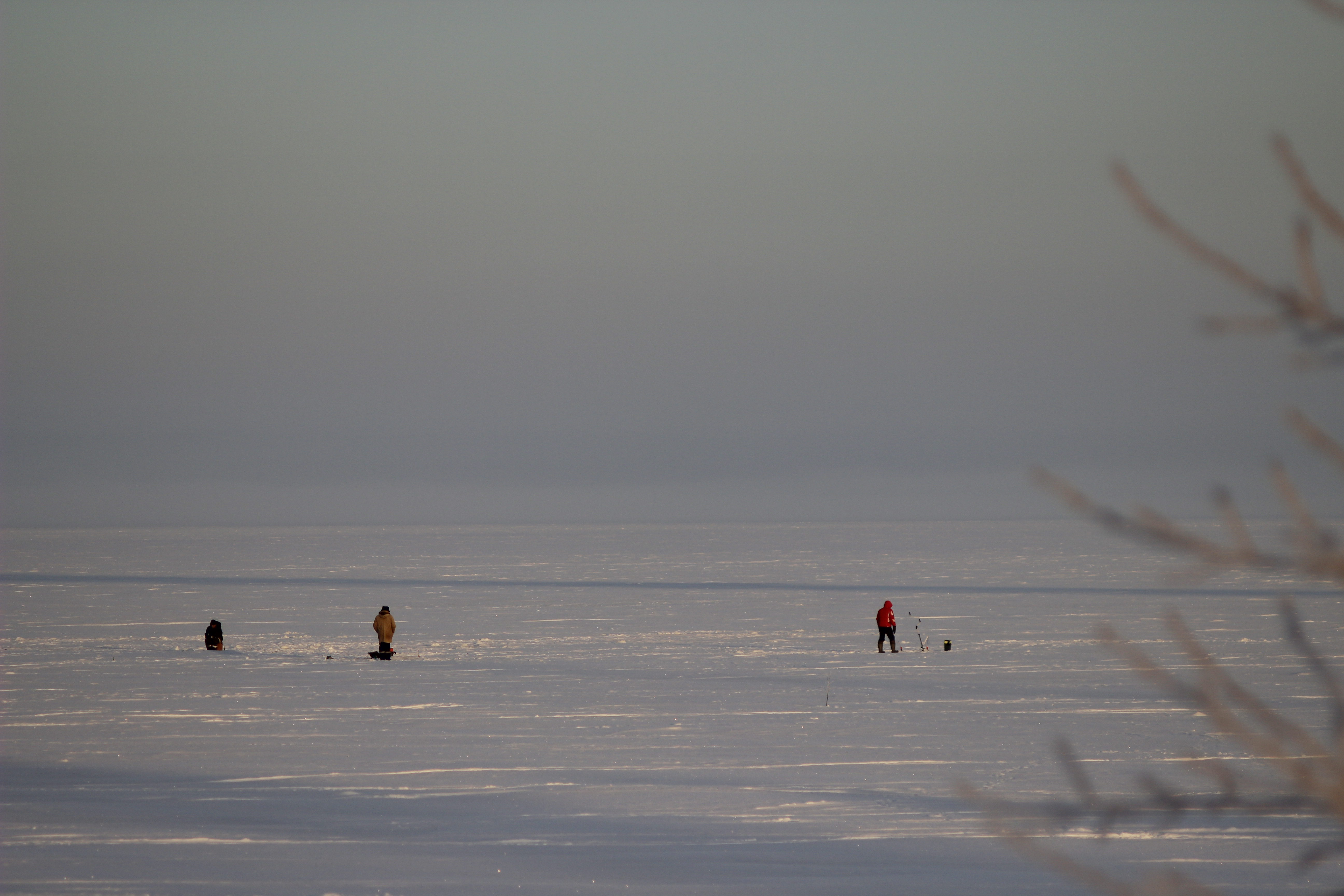Three people ice fishing on a vast frozen lake under a foggy winter sky.