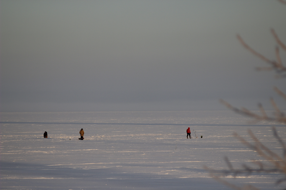Three people ice fishing on a vast frozen lake under a foggy winter sky.