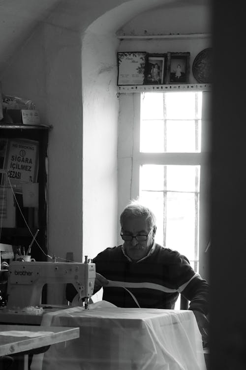 A Grayscale Photo of an Elderly Man Sitting Near the Sewing Machine