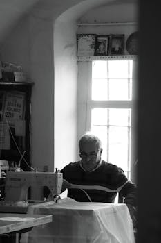 Monochrome image of an elderly tailor working at a sewing machine in a sunlit room.