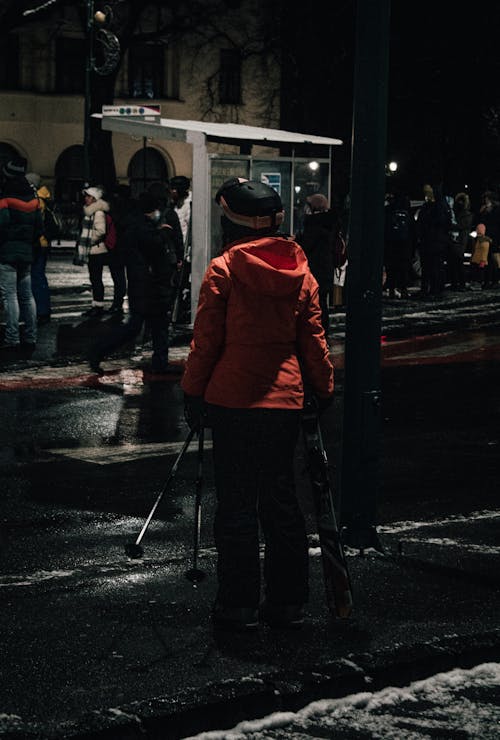 Free Person in Orange Jacket with Ski Poles Standing on the Street at Night Stock Photo