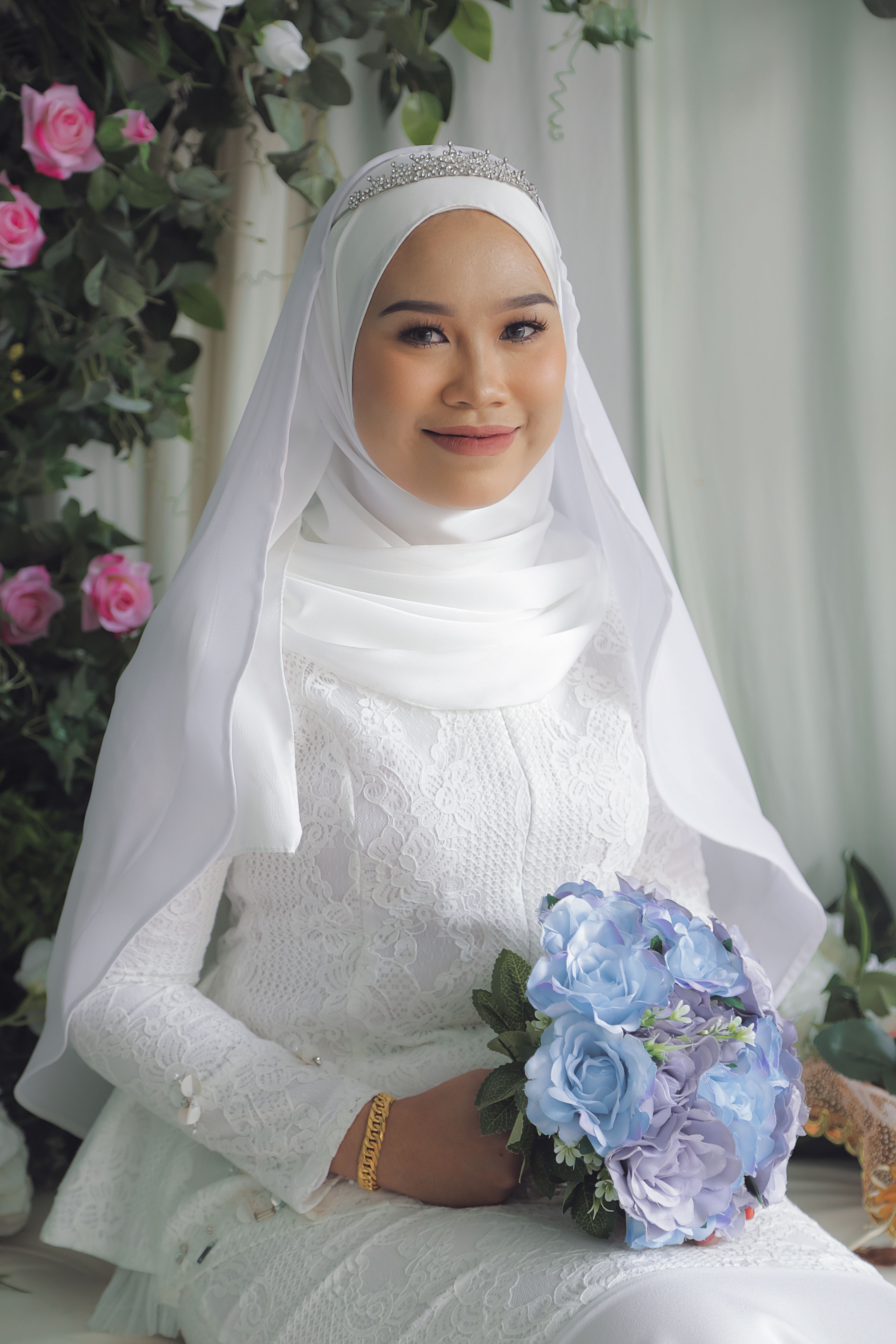 Portrait of a smiling Malay bride in a white wedding dress and hijab holding a bouquet.