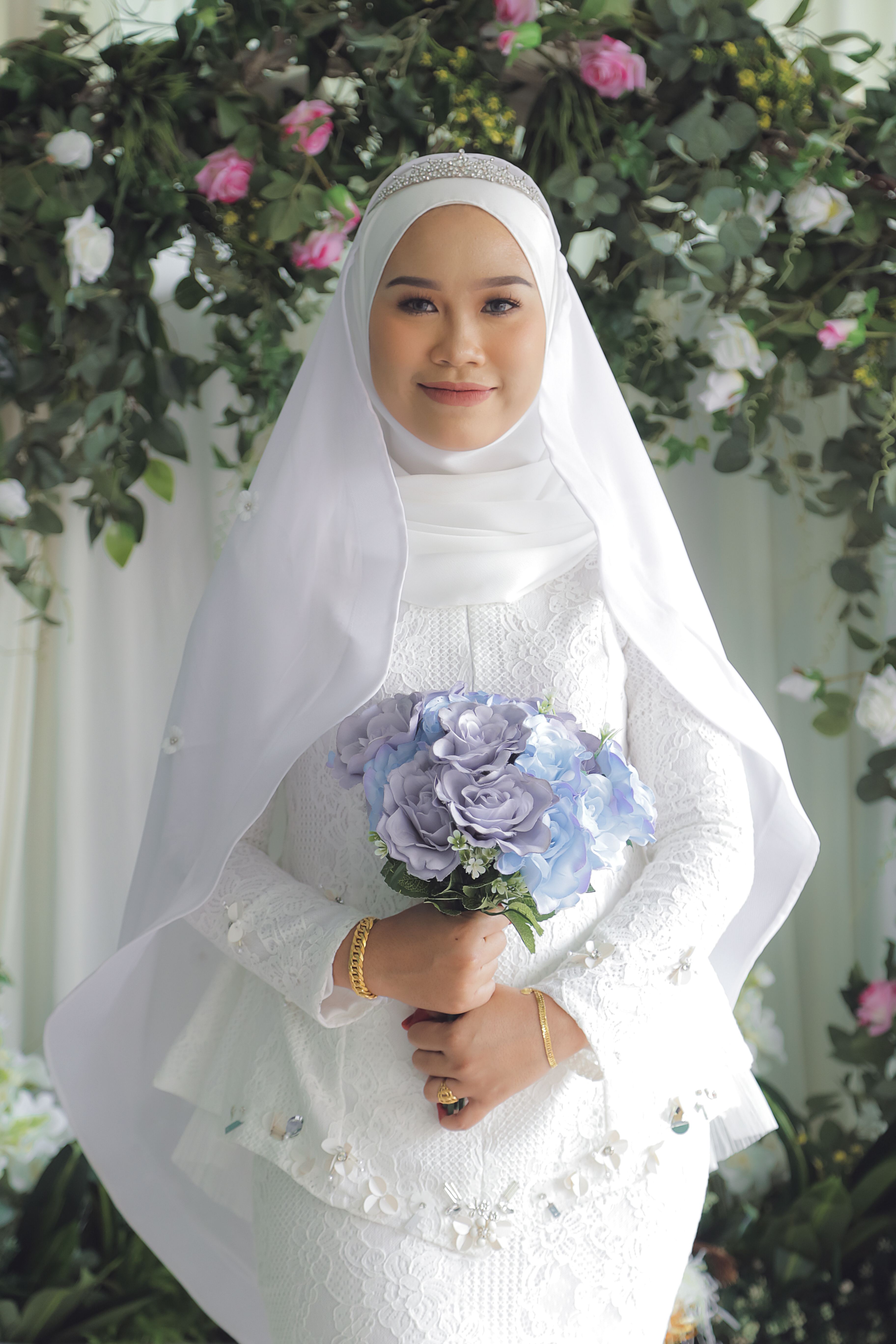 Beautiful Muslim bride in white gown holding a blue bouquet, smiling indoors.