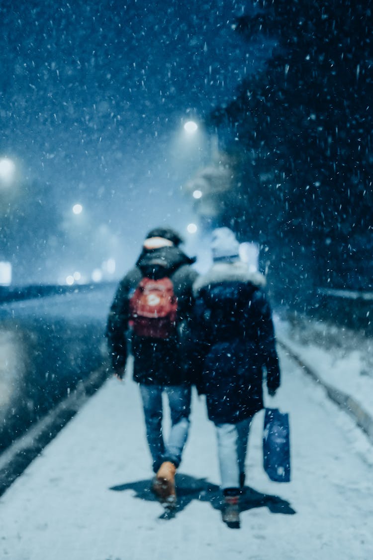 A Back View Of Two Persons Walking On A Snow Covered Ground At Night