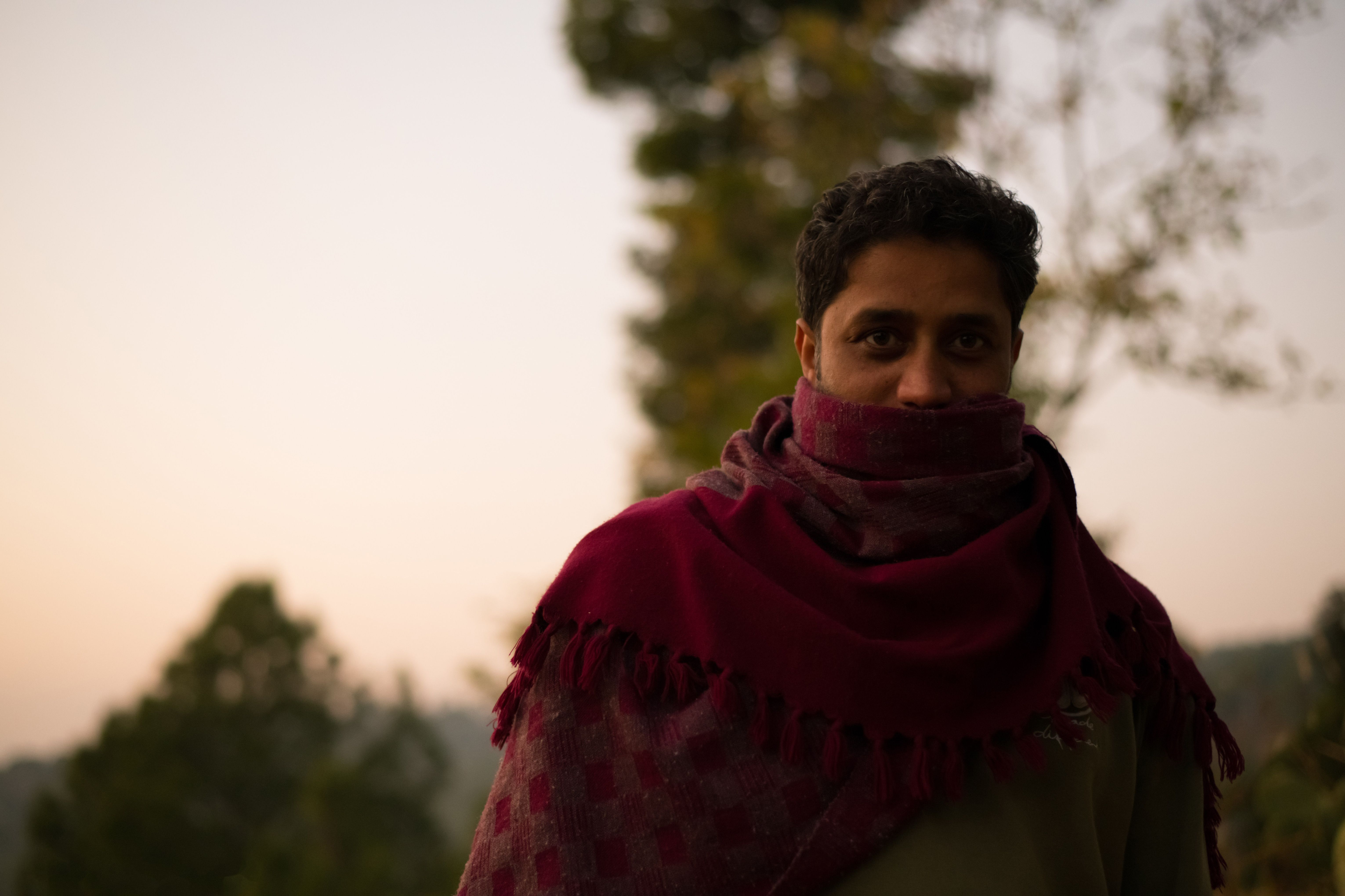 A man wrapped in a shawl stands outdoors at dusk, surrounded by trees.