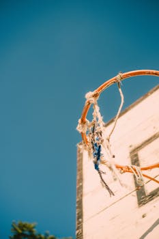 A low-angle view of a rustic basketball hoop with torn net under a clear blue sky.