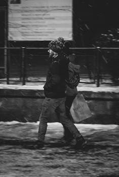 A couple walks through a snowy street at night, carrying bags and dressed warmly.