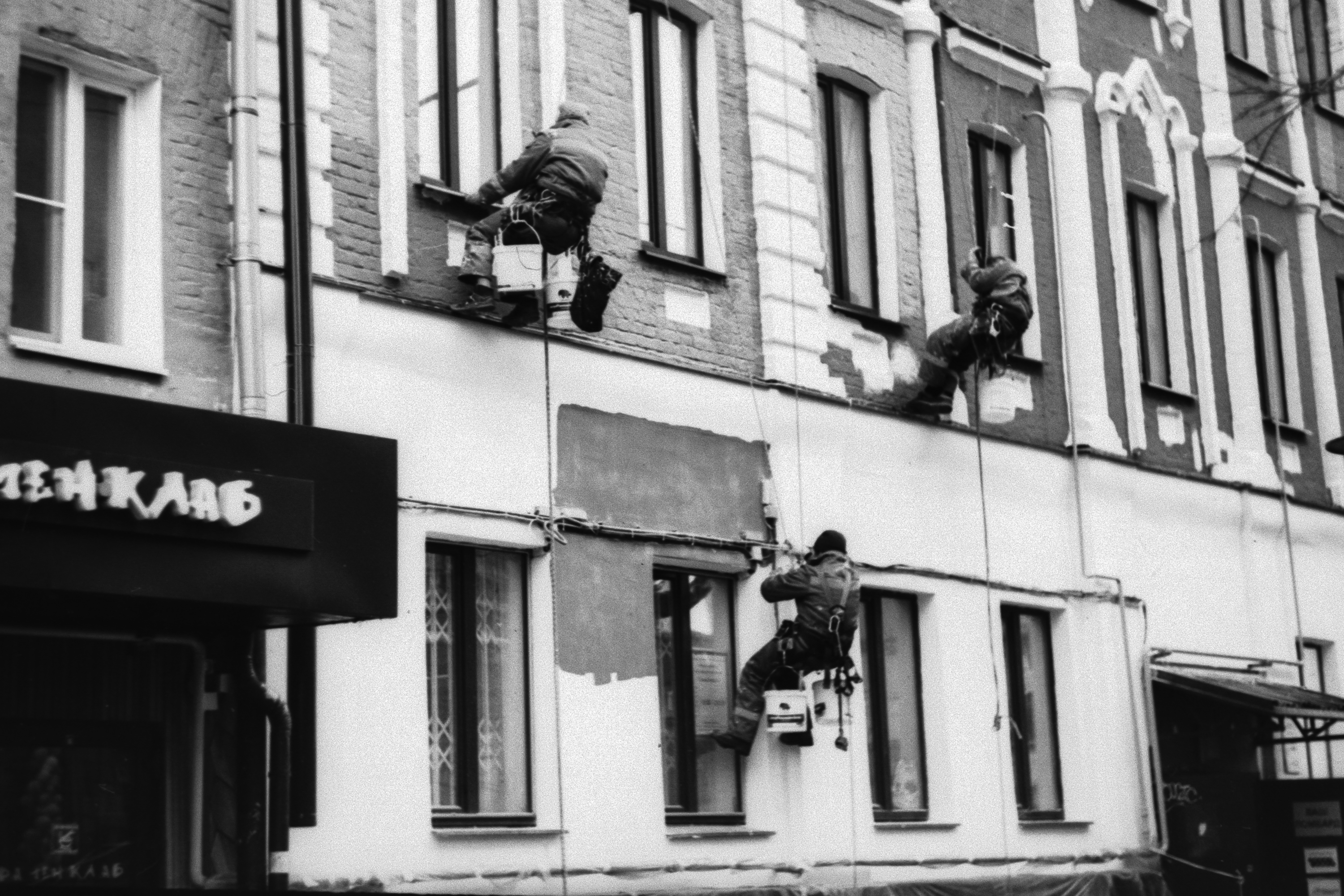 Three construction workers painting a building exterior, suspended by harnesses. Monochrome image.
