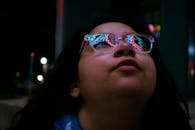 夜晚，一位年轻女子抬头，眼镜中反射出五颜六色的霓虹灯光
