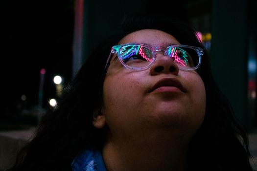 夜晚，一位年轻女子抬头，眼镜中反射出五颜六色的霓虹灯光