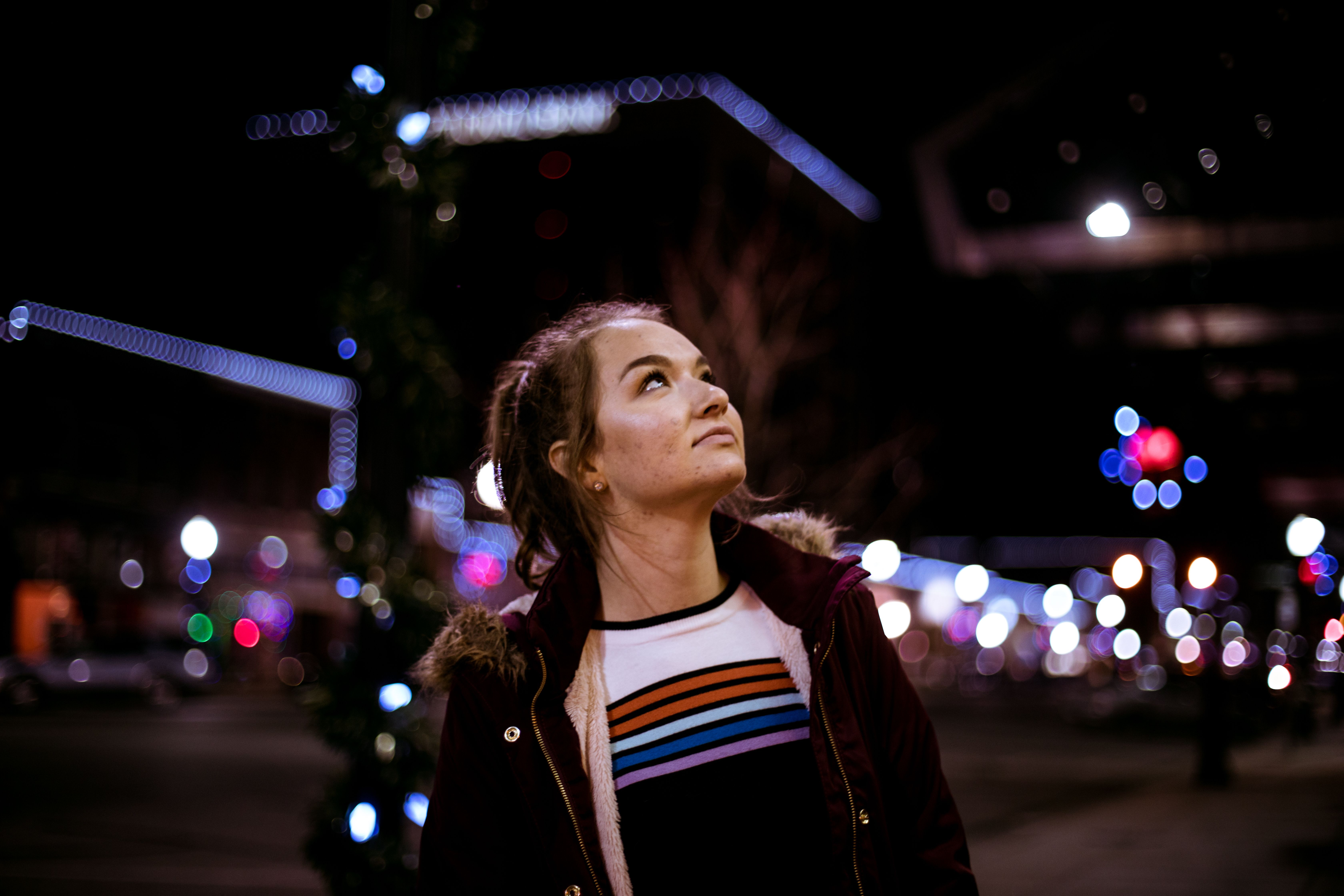 A woman in a jacket gazes upwards, surrounded by vibrant city lights at nighttime.