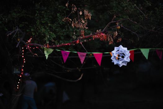 Colorful flags and paper decoration hanging from tree branches with string lights at nighttime.
