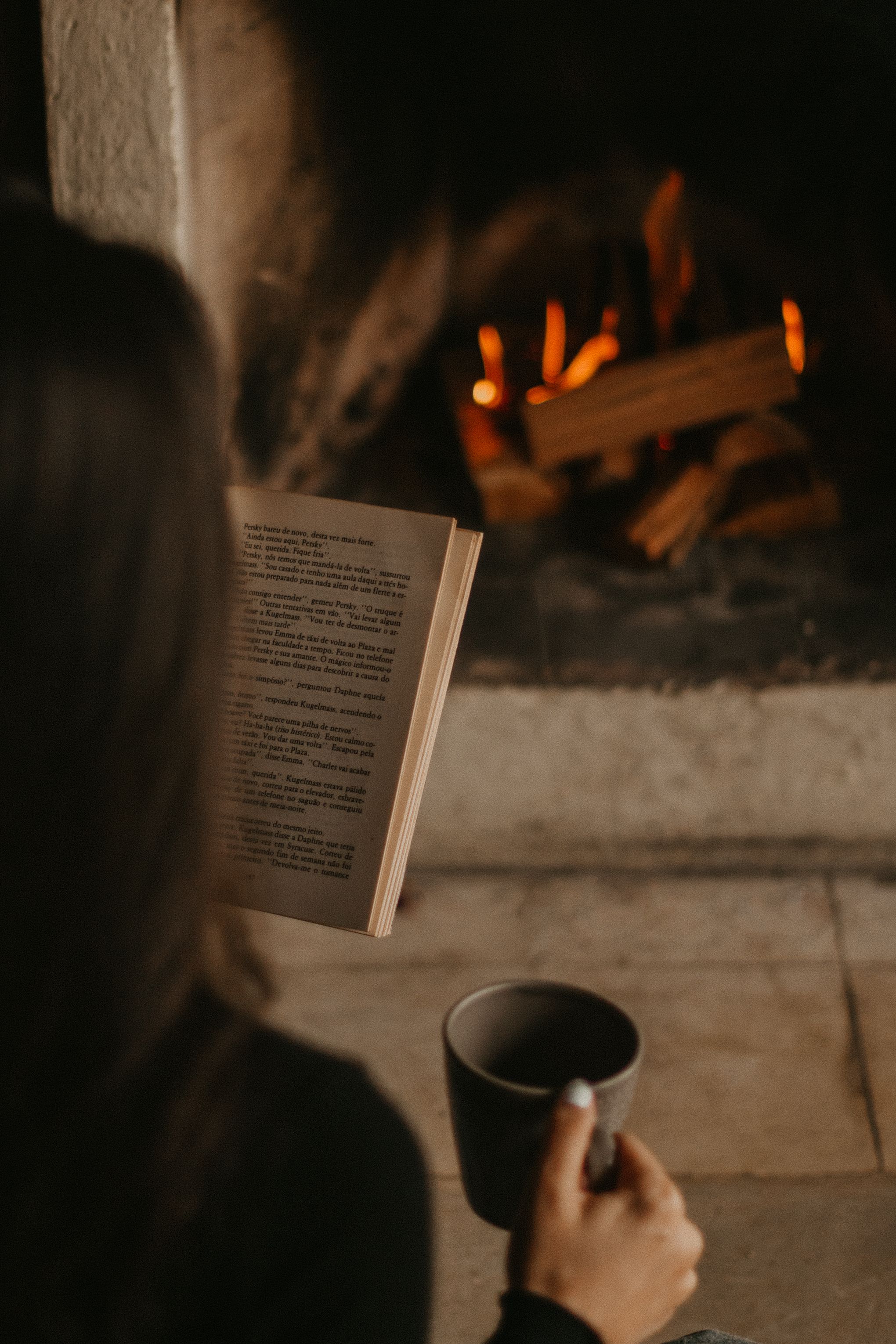 A woman enjoys reading by the fireplace, sipping coffee for a cozy evening ambiance.