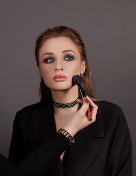 Portrait of a woman with professional makeup being applied, set against a neutral gray background.