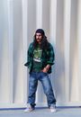 Man in Green and Black Camouflage Jacket and Blue Denim Jeans Standing Beside White Wall