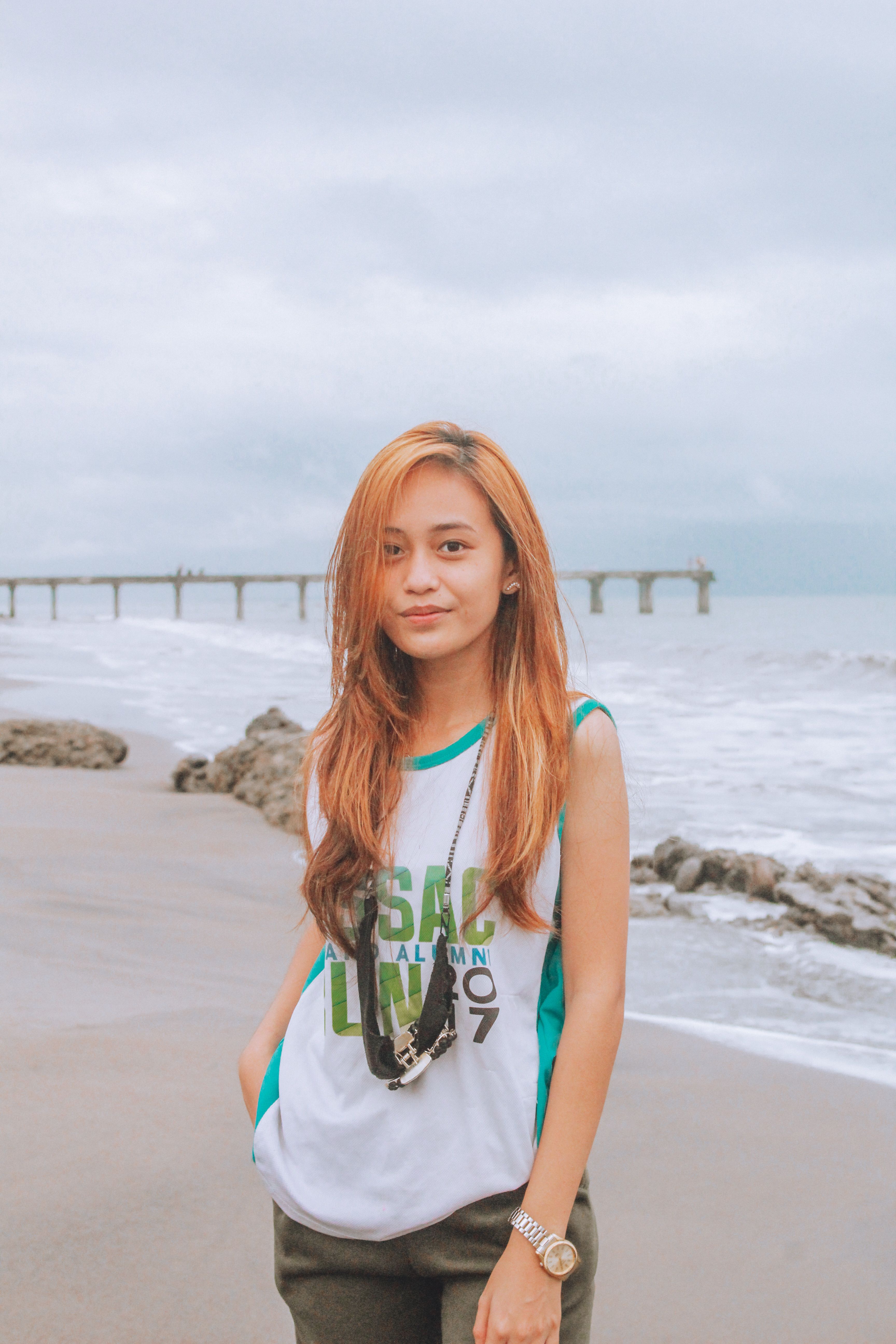 Portrait of a young woman standing on a sandy beach with a pier in the background on a cloudy day.
