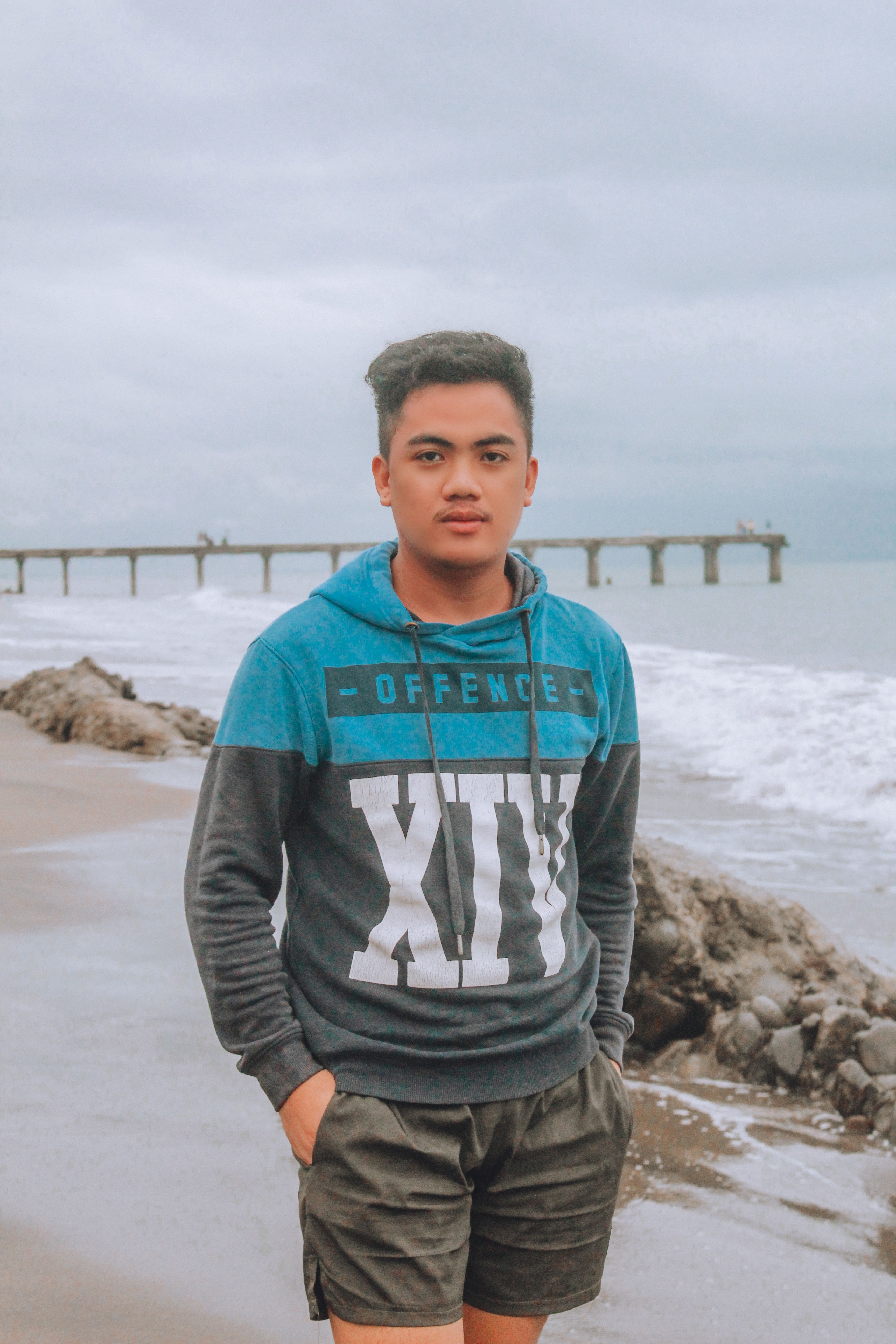 Young adult in casual wear standing on a sandy seashore with a pier in the background.