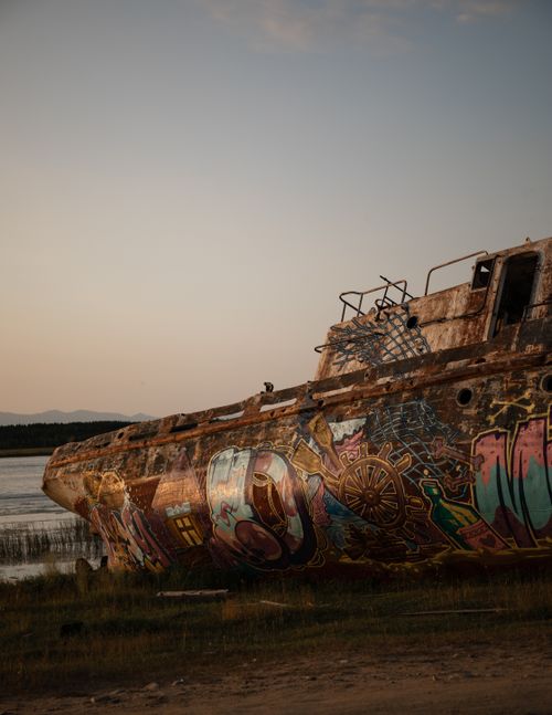 Free A colorful, graffiti-decorated shipwreck rests by a tranquil shoreline at dusk. Stock Photo