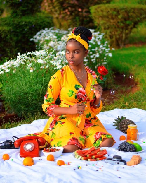 Free Elegant picnic setup with a woman in a floral dress holding a rose, surrounded by fruits and flowers, outdoors in Nairobi. Stock Photo