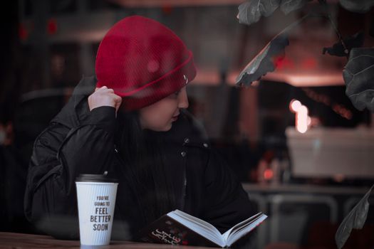 A stylish woman in a red beanie reads a book at a Tehran coffee shop, exuding a moody vibe.