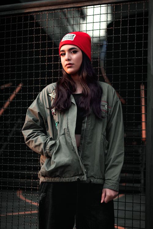 Free Portrait of a woman wearing a red beanie and green jacket, posing against a wire fence in an urban setting. Stock Photo