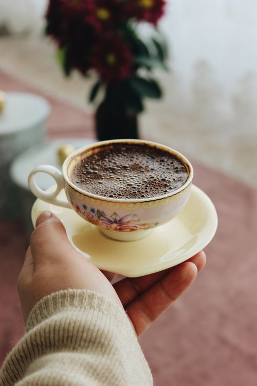 Free A close-up of a hand holding a floral teacup with coffee, set against a blurred background with flowers. Stock Photo