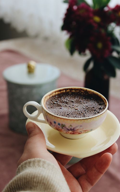 Free Close-up of a hand holding a foamy cup of hot chocolate in a floral ceramic cup with saucer. Stock Photo