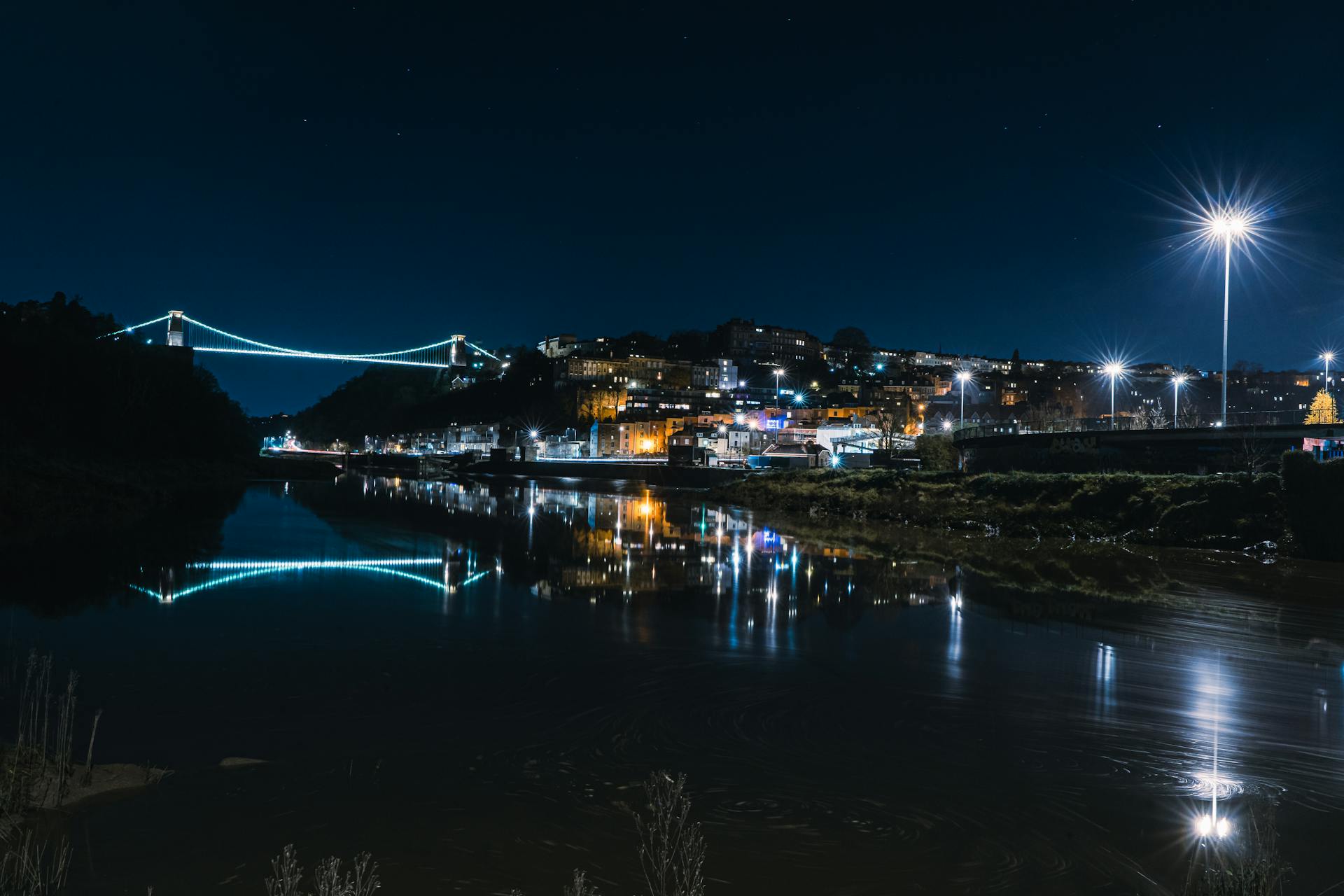 Scenic night view of Clifton Suspension Bridge reflecting over River Avon with city lights.
