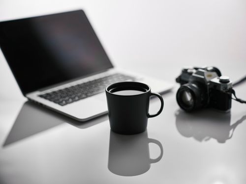 Free Minimalist workspace with a black mug, laptop, and DSLR camera on a reflective surface. Stock Photo
