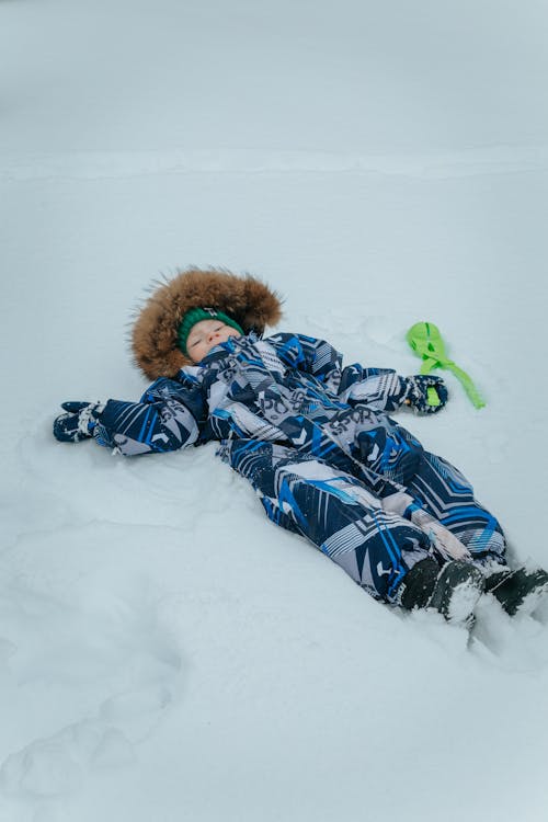 Free A Child in Blue and Gray Winter Clothes Lying on Snow Covered Ground Stock Photo