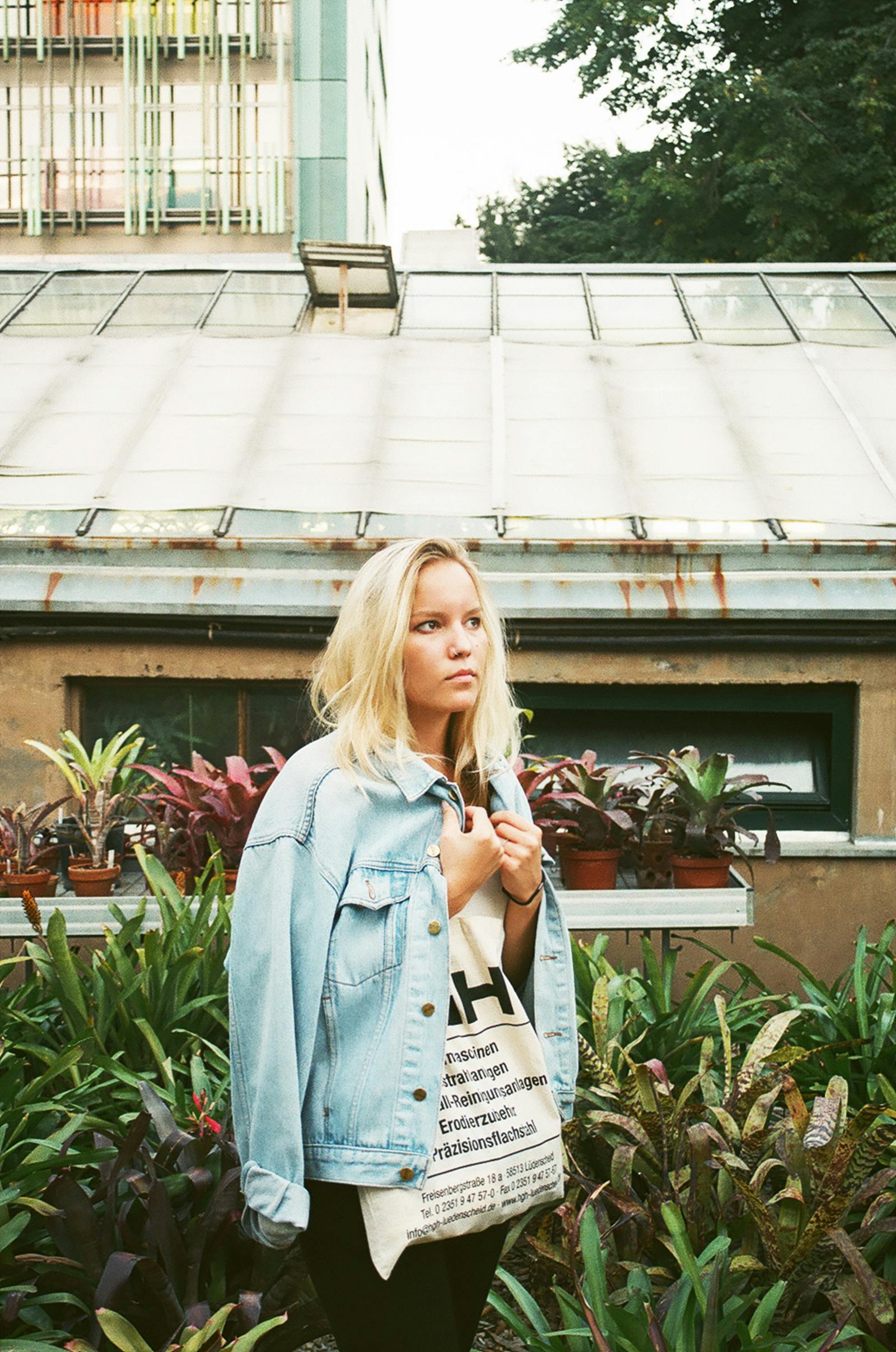 A young woman with blonde hair poses outdoors wearing a denim jacket, surrounded by urban greenery.