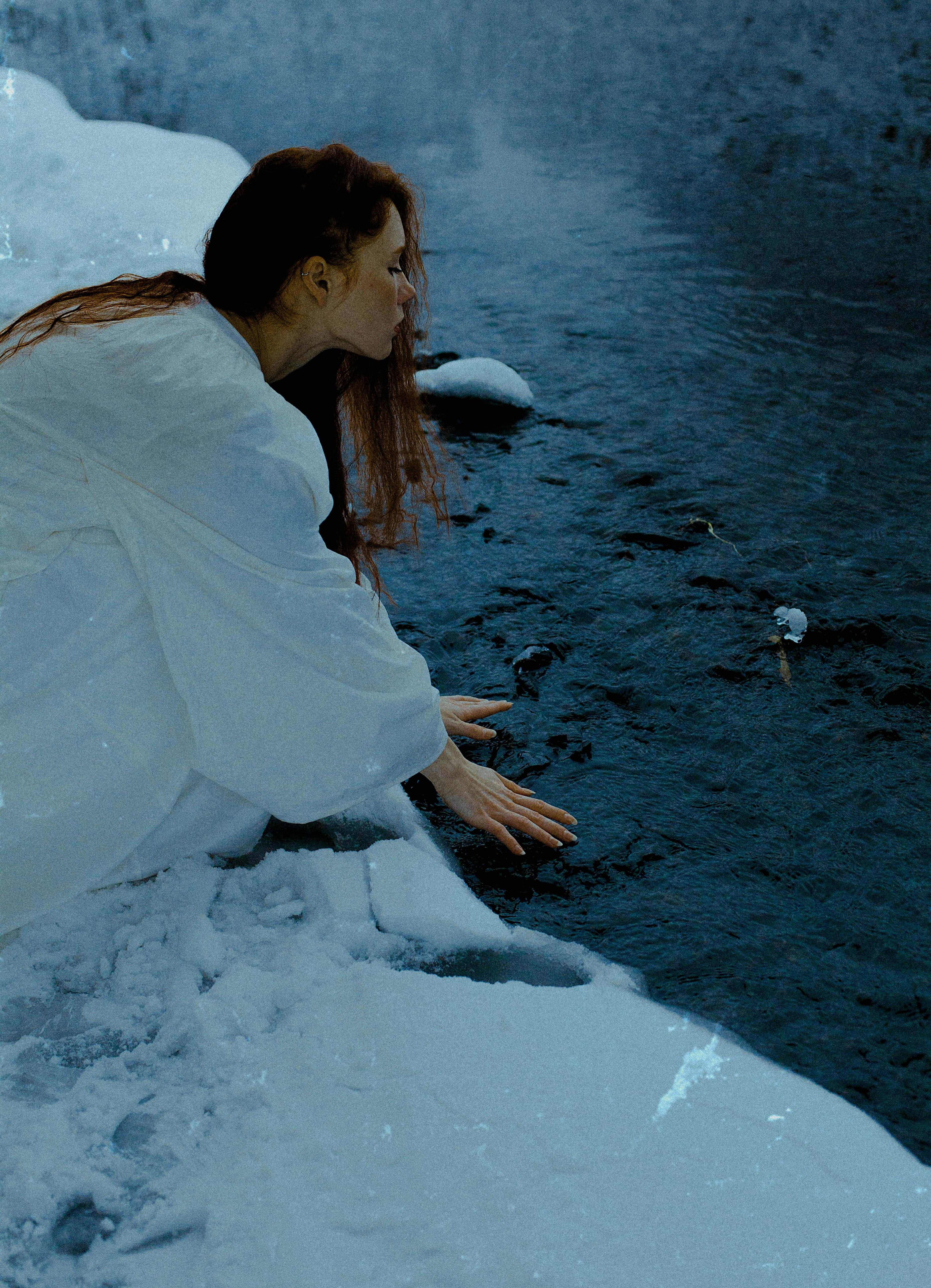 A woman in a white outfit reaches towards a snowy riverbank in Moscow during winter.