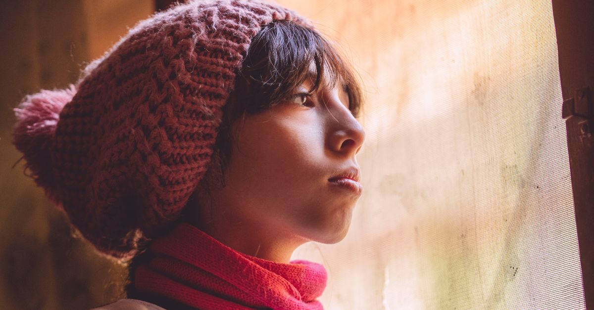 Teenager wearing winter knit cap gazes out a window, in warm indoor setting.