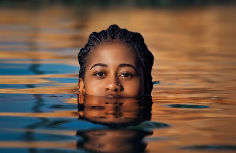 A Black woman with braided hair rests at the surface of calm water, her face above the waterline from the chin up, with orange and blue sunset light reflecting across the water around her.