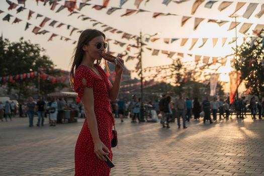 A fashionable woman enjoying a pretzel during sunset in bustling Istanbul's Taksim Square.