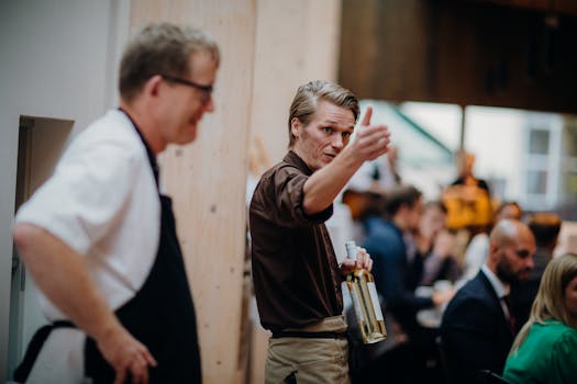 Waiter gesturing while holding wine bottles at a lively restaurant setting.