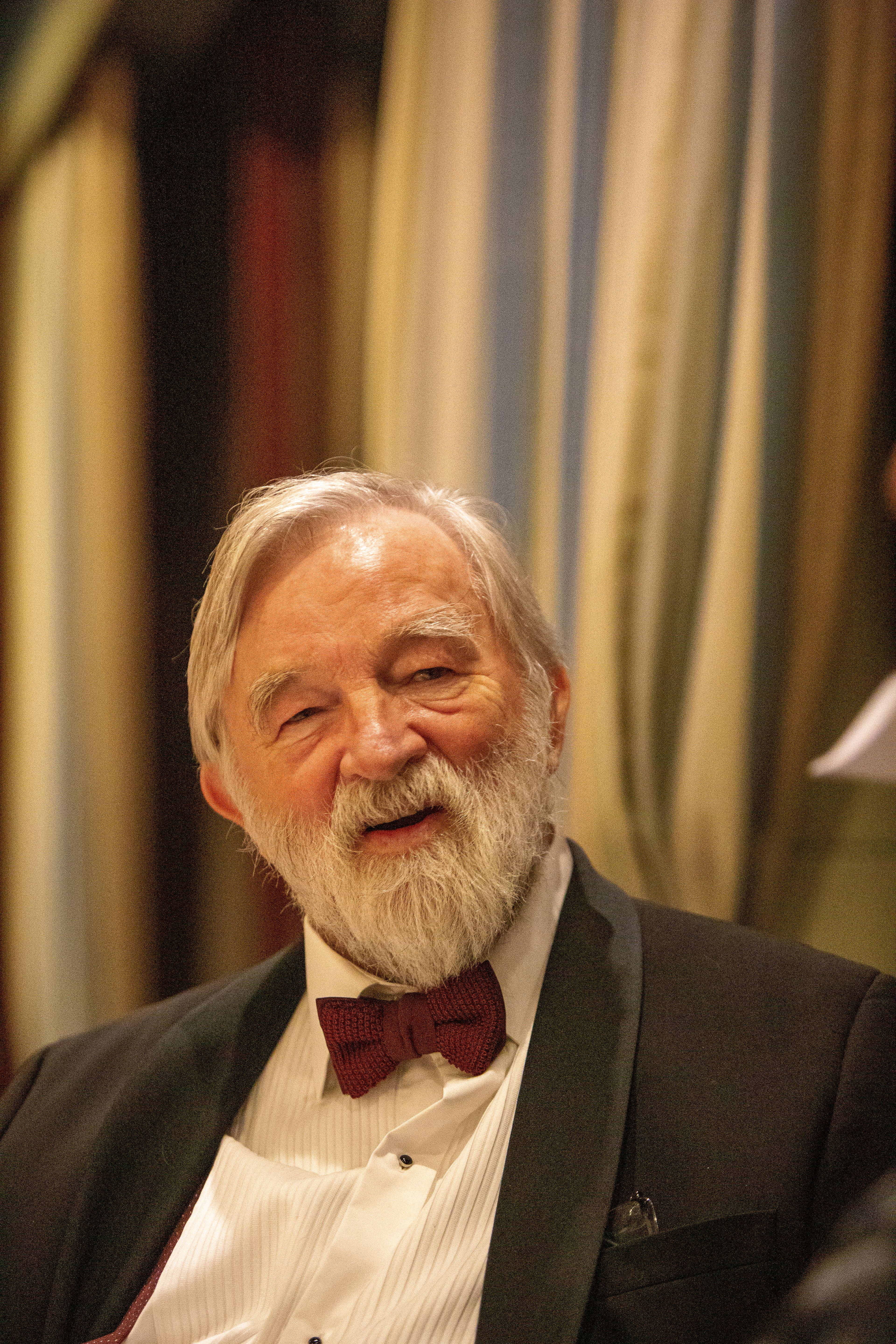 Senior man with a beard in a black tuxedo and bowtie smiling warmly indoors.