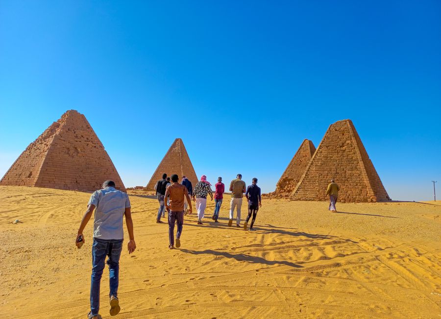 Tourists walk towards ancient pyramids in the Sudanese desert under a clear blue sky.