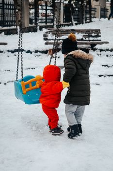 Two children in winter clothing enjoy a snowy playground swing.