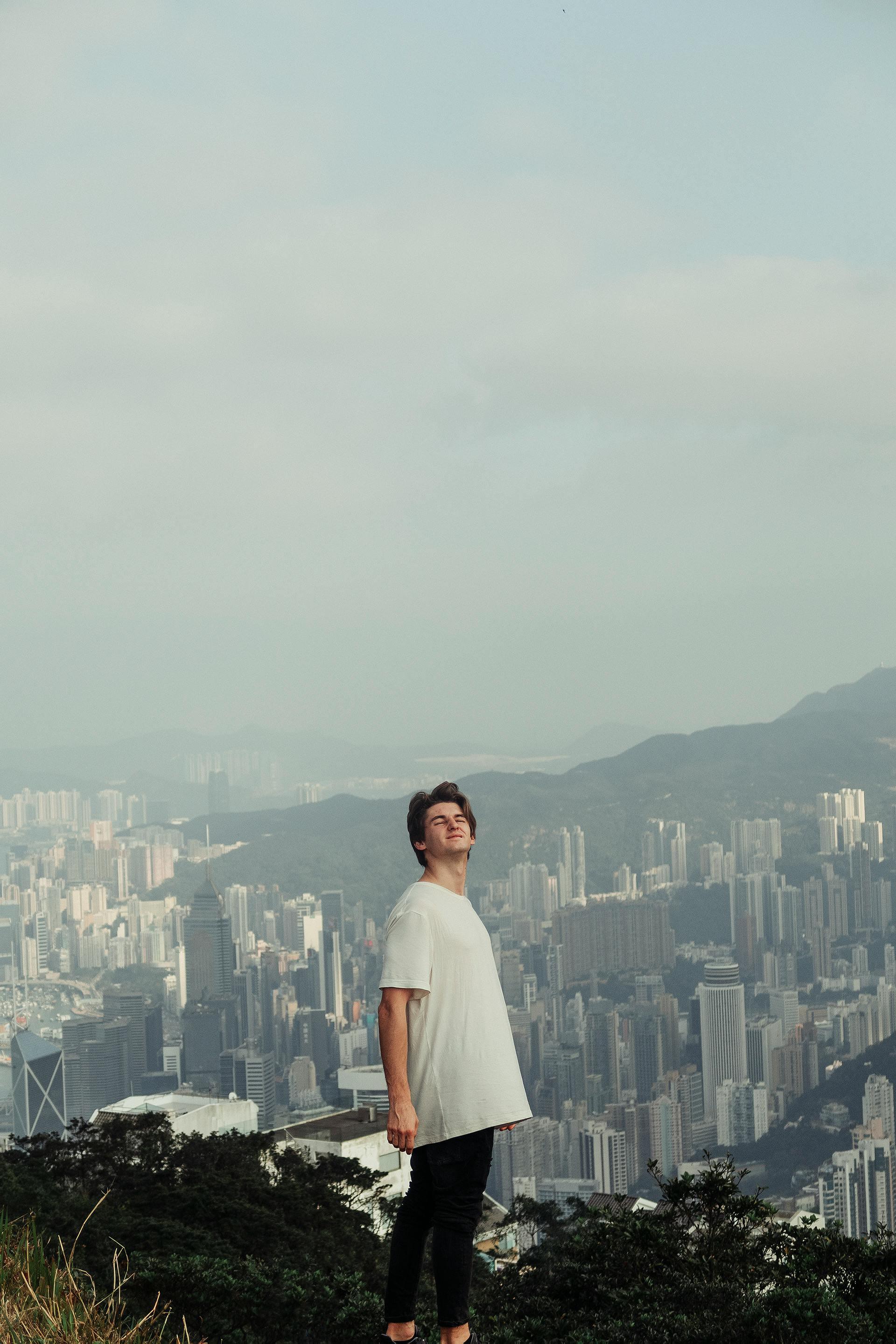A man standing on a mountain top overlooking the Hong Kong skyline on a clear day.