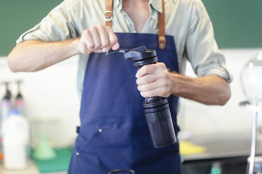 A barista wearing an apron grinds coffee beans manually using a hand grinder inside a cafe.