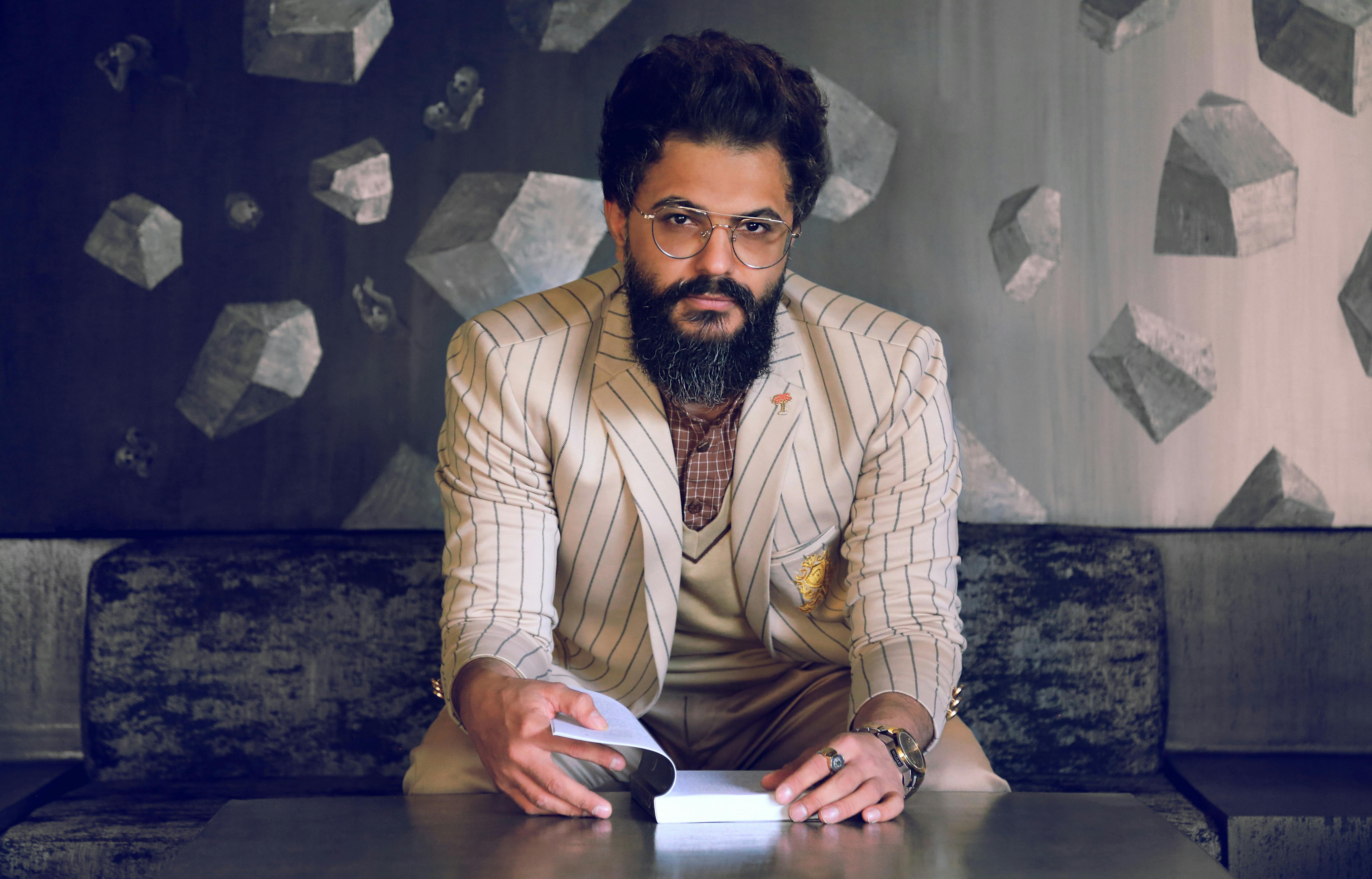 Portrait of a stylish bearded man in elegant attire, seated indoors with eyeglasses, exuding confidence.