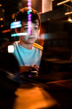 Woman working on a laptop with colorful reflections visible on screen in a dimly lit room.