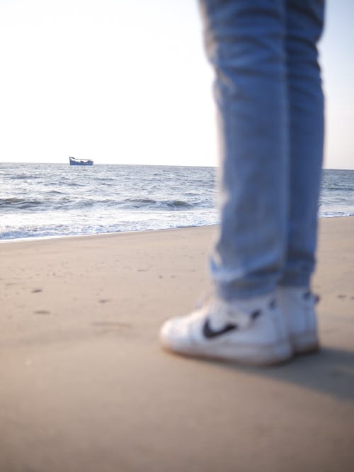 Person in Blue Denim Jeans and White Sneakers Standing on the Beach