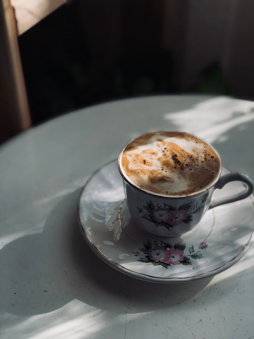 Free Close-up of a frothy cappuccino in a floral ceramic cup on a sunlit table. Stock Photo