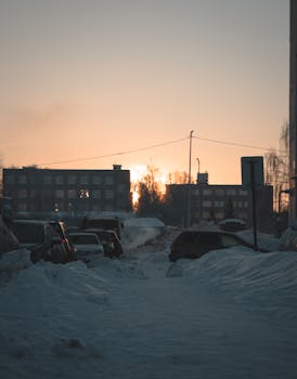 A serene winter cityscape with cars parked on a snow-covered street during sunset.
